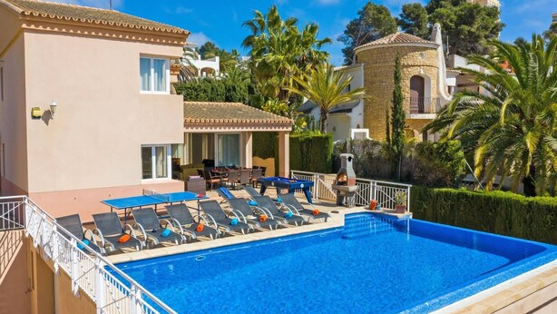 Área de piscina com espreguiçadeiras, churrasqueira e um edifício com telhado de azulejos.