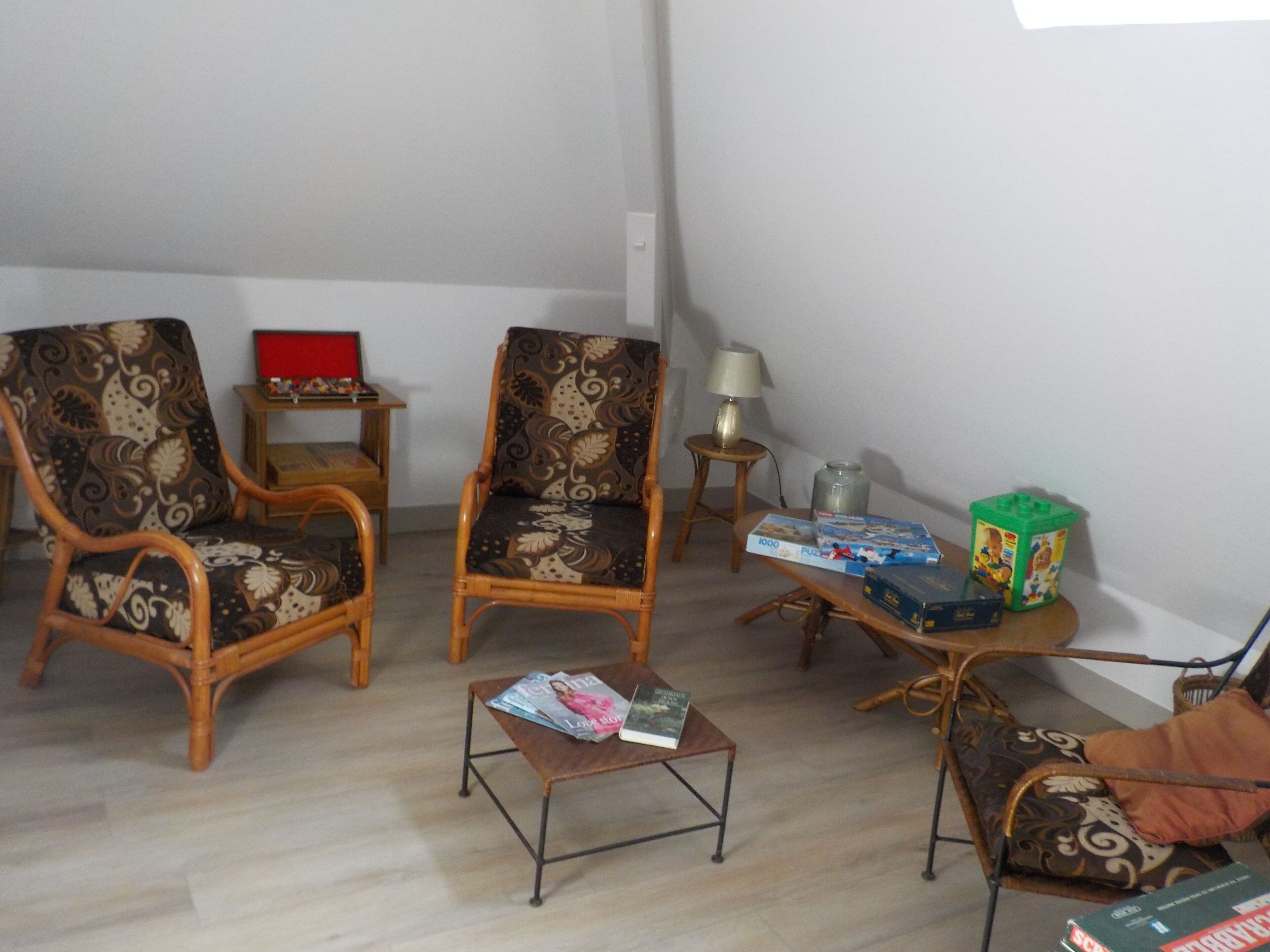 A living room with two patterned armchairs, a small coffee table with magazines, a side table with a lamp, and a green toy box.