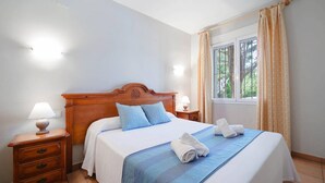 Ein ordentlich bezogenes Bett mit blauer Bettdecke und Kissen, zwei Nachttischlampen, ein Holznachttisch und ein Fenster mit Vorhängen.