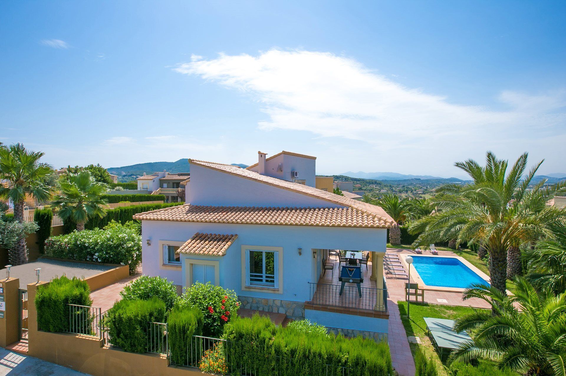 A villa with a swimming pool, surrounded by lush greenery and palm trees, overlooking a scenic landscape.