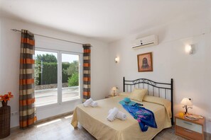 A bedroom with a bed, a nightstand, a lamp, a painting, and a window with curtains. - Villa Cabriel by Villa Plus (Jávea)
