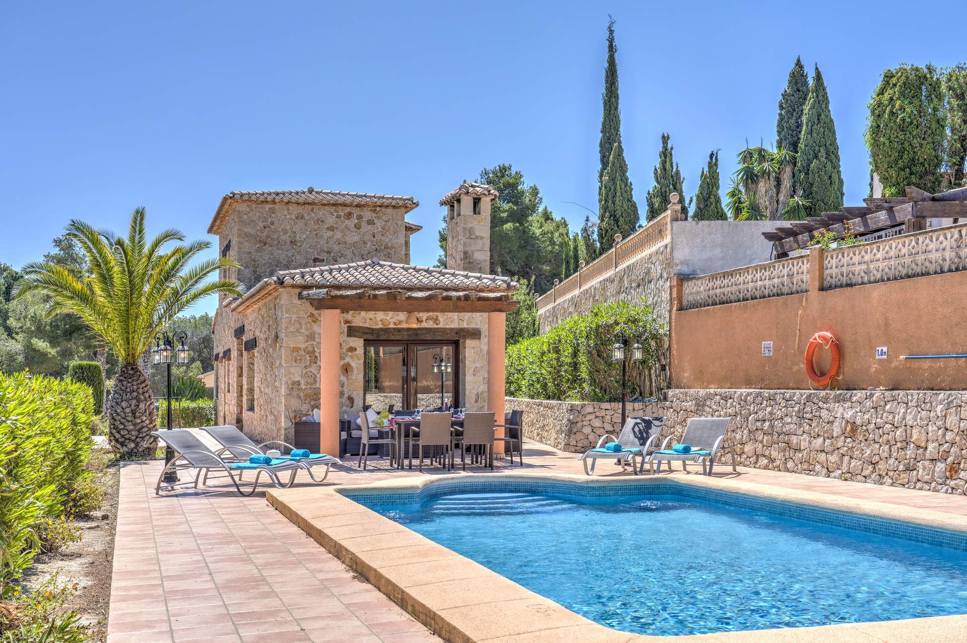 A pool area with lounge chairs, a small building with a tiled roof, and a stone wall.
