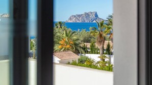 A view of a mountainous landscape with a clear blue sea, palm trees, and a white building with a tiled roof. - Villa Catorce Flores by Villa Plus (Teulada)