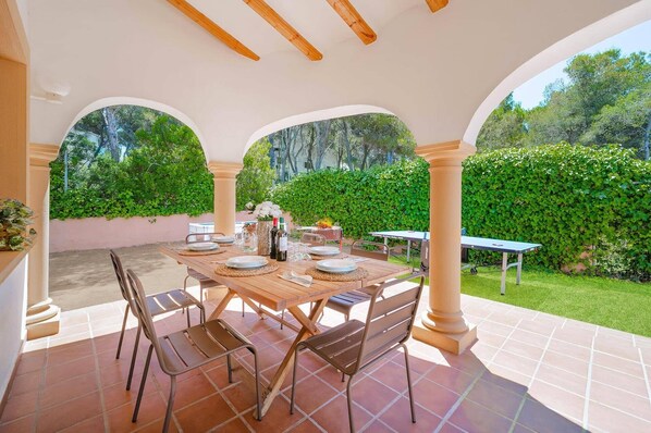 An outdoor patio with a table set for dining, surrounded by chairs, and a view of a lush garden.