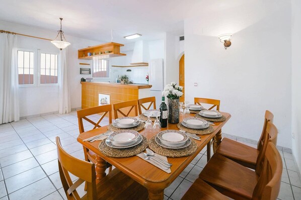 A dining area with a wooden table set for six, surrounded by chairs. A kitchen area is visible in the background with a counter and cabinets.