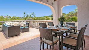 Um pátio externo com móveis de vime, uma mesa posta para refeições e vista para a paisagem.