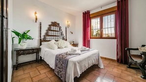 Quarto com cabeceira de madeira, mesa de cabeceira com uma planta, janela com cortinas vermelhas e uma cadeira.