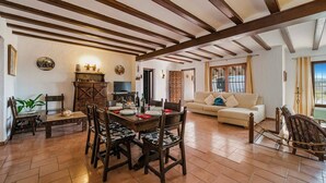 Uma sala de jantar com uma mesa e cadeiras de madeira, televisão e sofá, em um ambiente com vigas de madeira aparentes e piso ladrilhado.