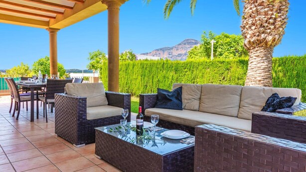 An outdoor patio with wicker furniture, a glass-top table, and a view of a mountain and greenery. - Villa Elefante by Villa Plus (Jávea)