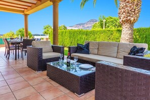 Eine Terrasse im Freien mit Korbmöbeln, einem Tisch mit Glasplatte und Blick auf Berge und Grünpflanzen.