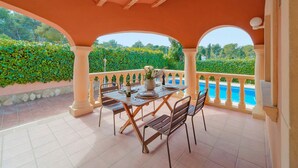 A covered outdoor dining area with a table set for two, overlooking a swimming pool and lush greenery. - Villa Fresa Blanca by Villa Plus (El Tosalet)