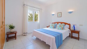 A bedroom with a bed, two nightstands, a window with curtains, a small table with a plant, and wall-mounted lights. - Villa Fresa Blanca by Villa Plus (El Tosalet)