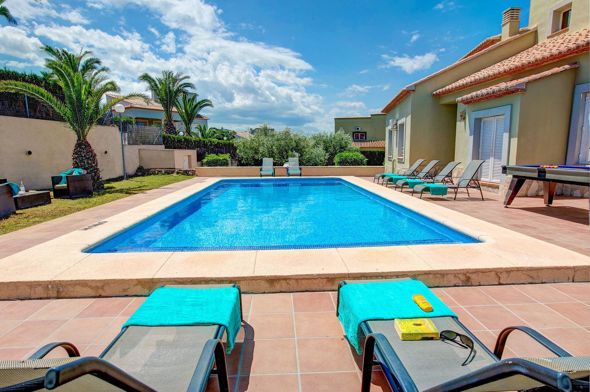 A swimming pool with lounge chairs and a sunbed in the foreground, surrounded by a landscaped garden and a building with a tiled roof.