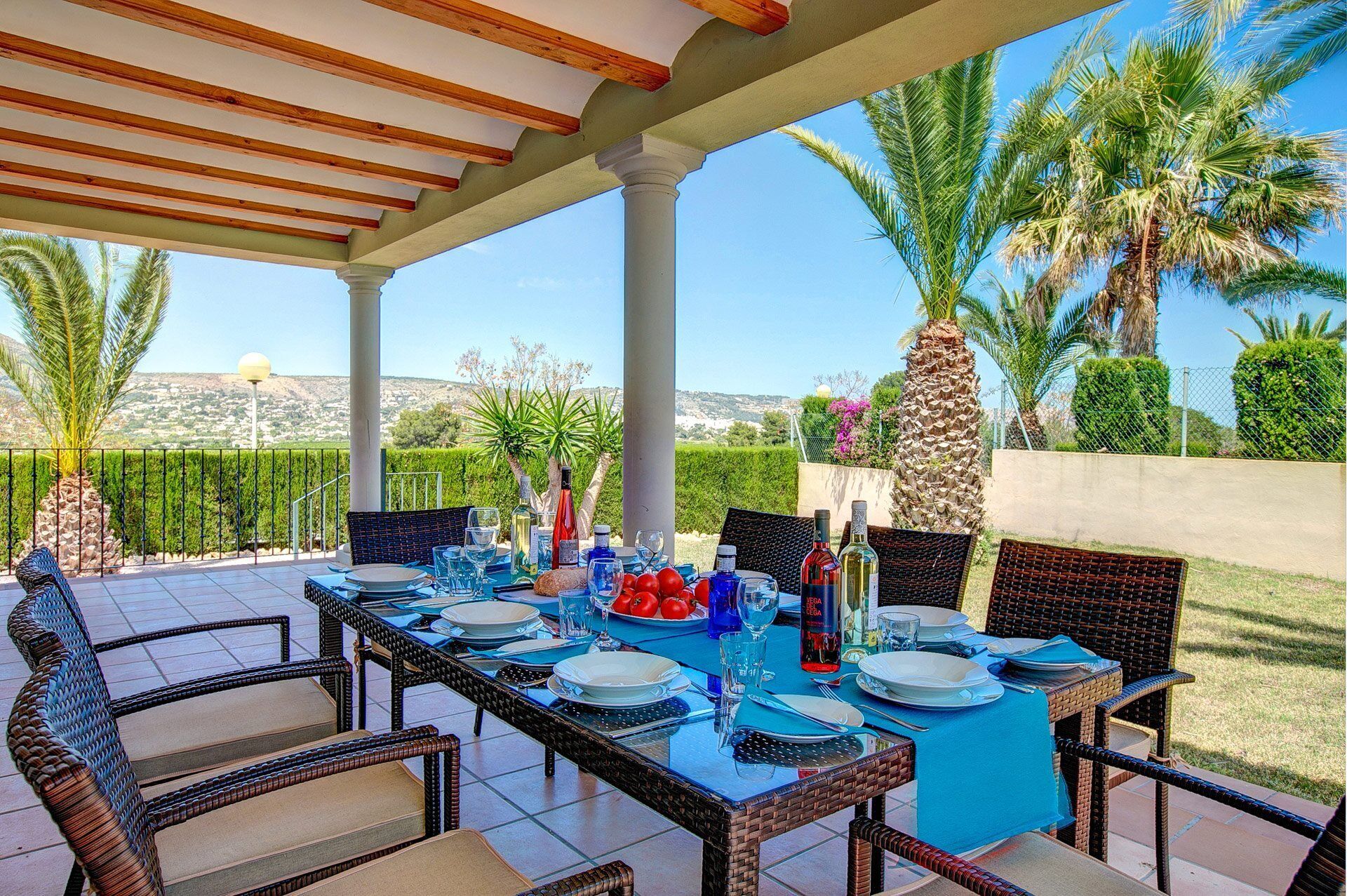 An outdoor dining area with a covered pergola, wicker chairs, and a table set for a meal with blue tablecloths.