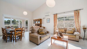 Ein geräumiges Wohnzimmer mit Essbereich, ausgestattet mit einer Sofagarnitur, einem Couchtisch und einem Esstisch mit Stühlen. Es gibt ein großes Fenster mit Vorhängen, eine Klimaanlage und einen Schrank mit Dekoration.
