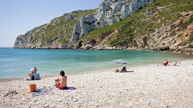 Ein Kiesstrand mit badenden Personen und einer felsigen Klippe im Hintergrund.
