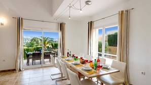 Un espace repas avec une longue table dressée pour six personnes, de grandes fenêtres avec des rideaux et une vue sur un paysage verdoyant.