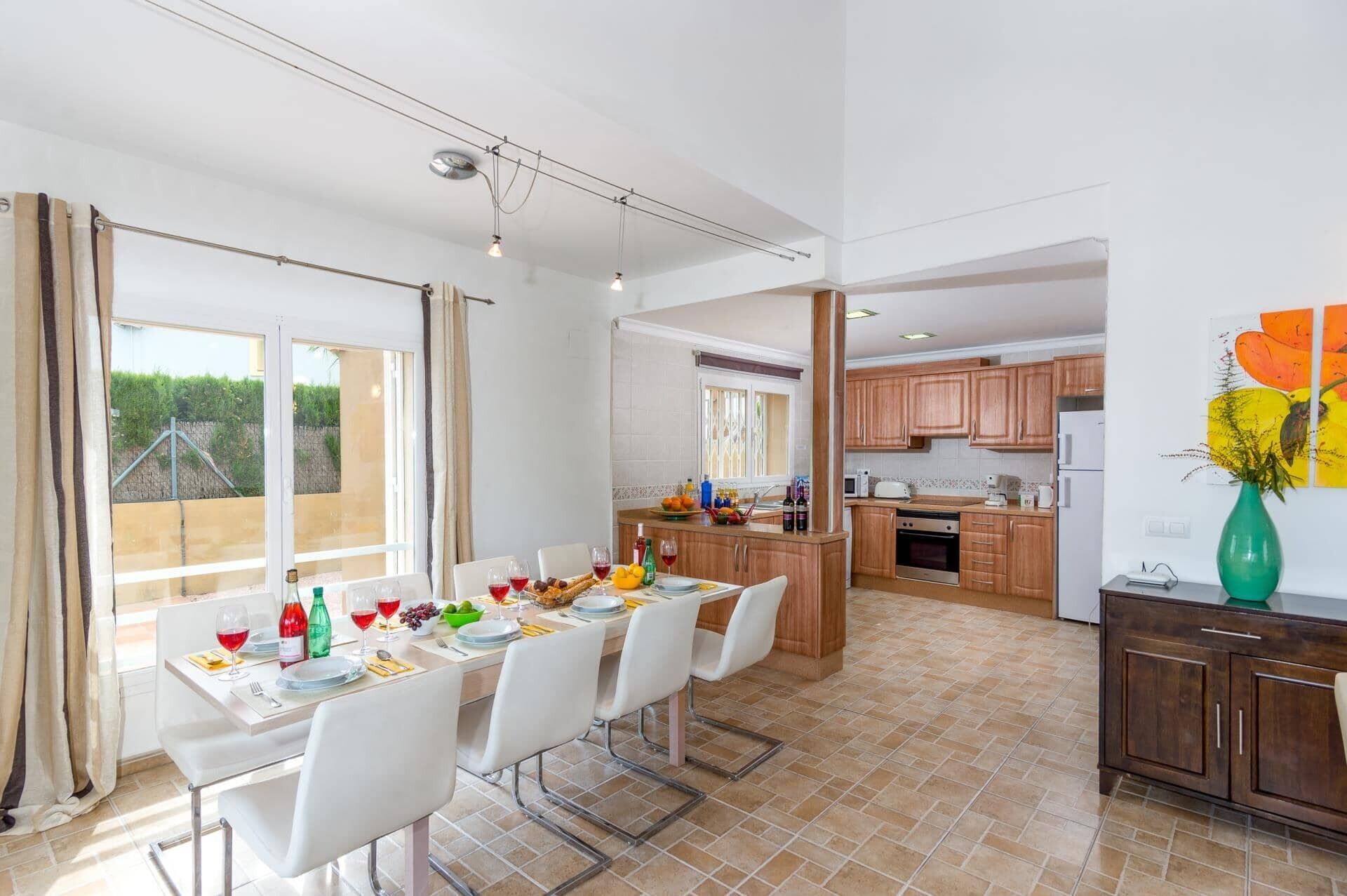 A spacious dining area with a long table set for six, a kitchen with wooden cabinets, and a view of greenery outside.