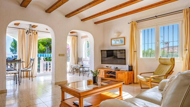 A spacious living room with a wooden coffee table, a sofa, and a TV. There are two windows with curtains, a balcony visible through an archway, and a dining area with a table and chairs. - Villa Moggalatto by Villa Plus (Calp)