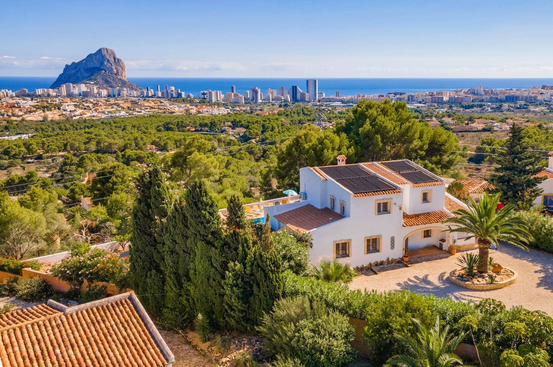 Une villa blanche, dotée d’un toit en tuiles, entourée d’une végétation luxuriante et offrant une vue sur un paysage urbain, avec une montagne imposante qui se profile à l’horizon.