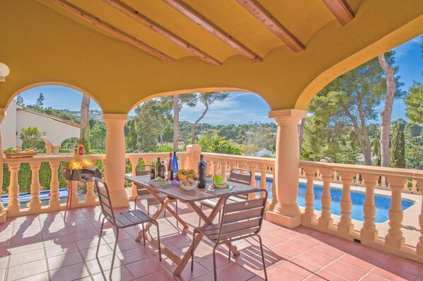 A covered patio with a table and chairs, a grill, and a view of a swimming pool and greenery.
