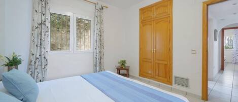 A bedroom with a bed, curtains, a wooden wardrobe, a small table with a plant, and a window with a view of trees.