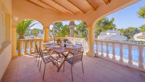 Un área de comedor al aire libre techada con una mesa preparada para cuatro personas, rodeada de sillas, con vista a una piscina y palmeras.