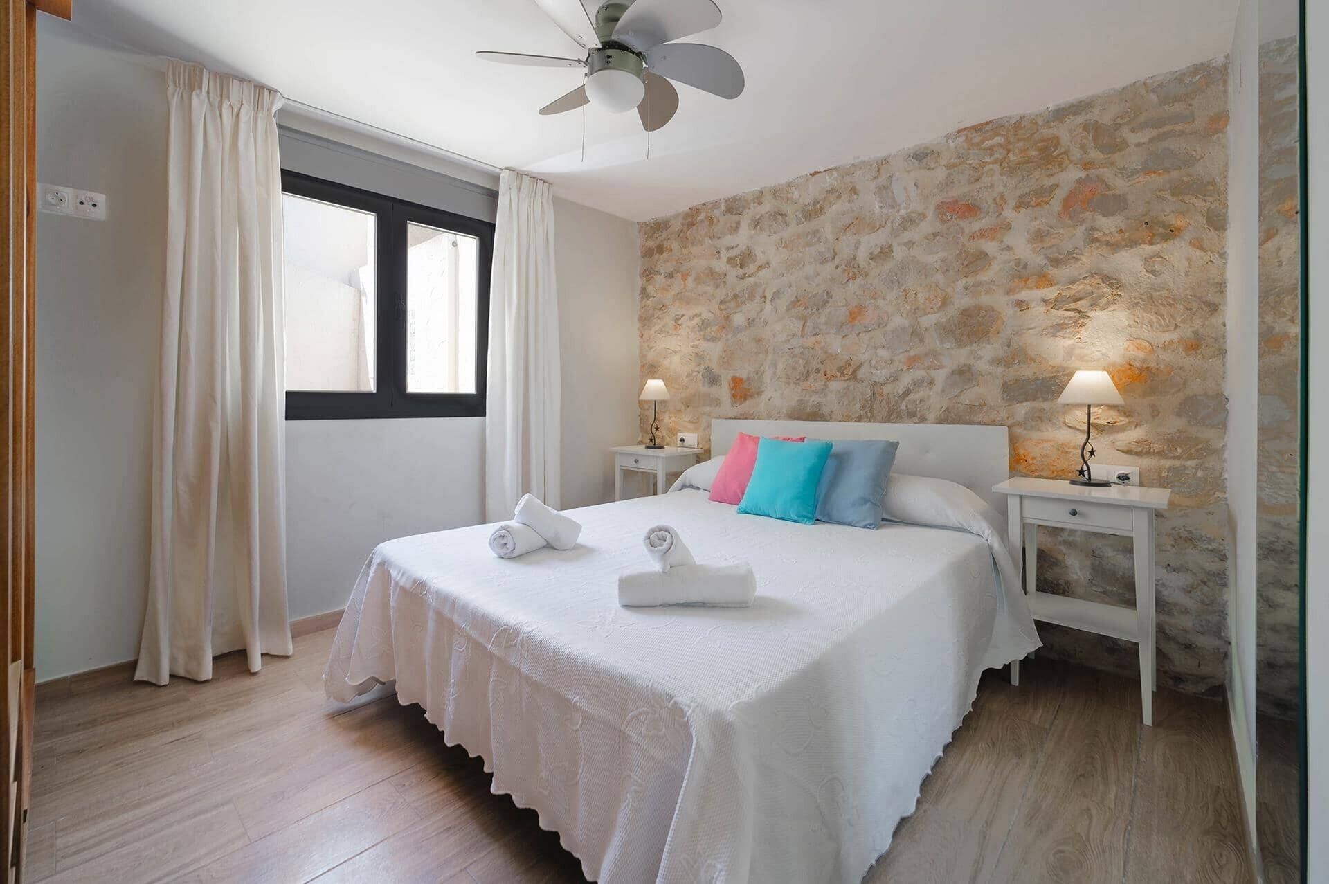 Une chambre avec un mur en pierre, un lit avec une literie blanche et deux oreillers, un ventilateur de plafond et deux lampes de chevet.