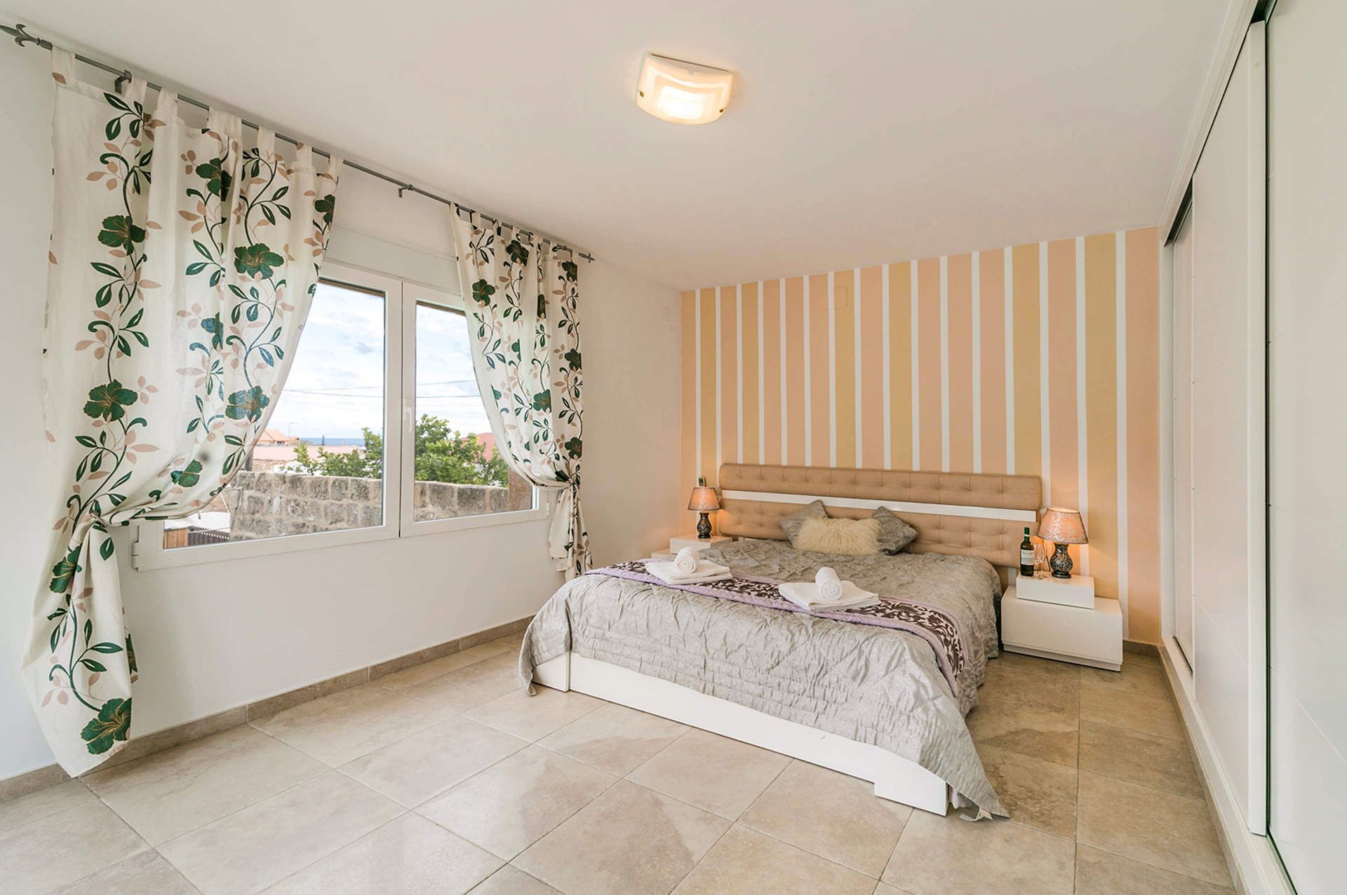 A bedroom with a bed, two bedside tables, a window with curtains, a striped wall, and a ceiling light.