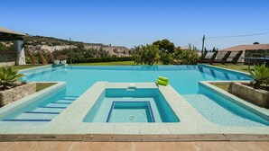 En rektangulär swimmingpool med en central del avsedd för dykning, omgiven av en terrass med solstolar och en liten trädgård.