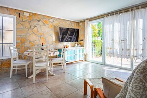 A dining area with a stone wall, a TV mounted above a table, and a view of greenery through large windows. - Villa Silvasol by Villa Plus (Javea)