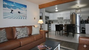 A living room with a brown leather sofa, a coffee table with a magazine, and a painting of a snowy landscape. - Renovated Condo steps from the Beaver Creek shuttle! (Avon)