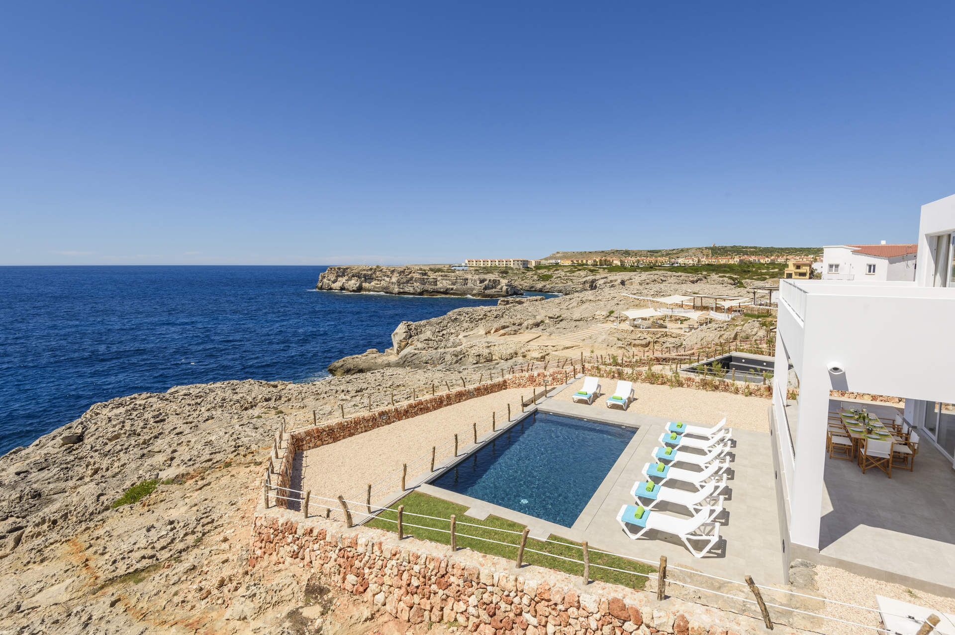 A pool area with lounge chairs, a clear view of the sea, and rocky terrain.