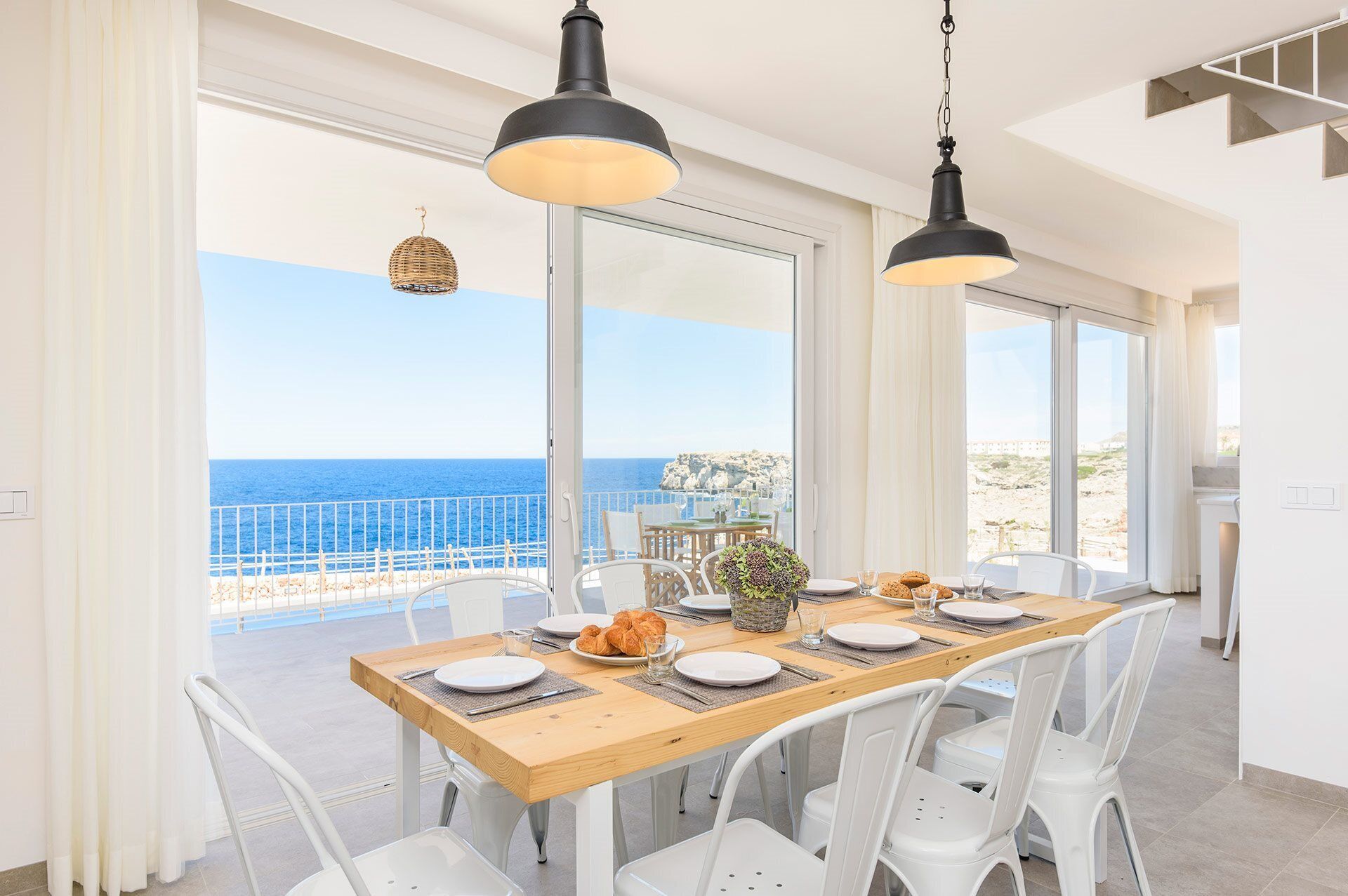 Un comedor con una mesa de madera dispuesta para cuatro personas, sillas blancas y vistas al mar a través de amplios ventanales.