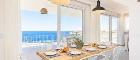Un comedor con una mesa de madera dispuesta para cuatro personas, sillas blancas y vistas al mar a través de amplios ventanales.
