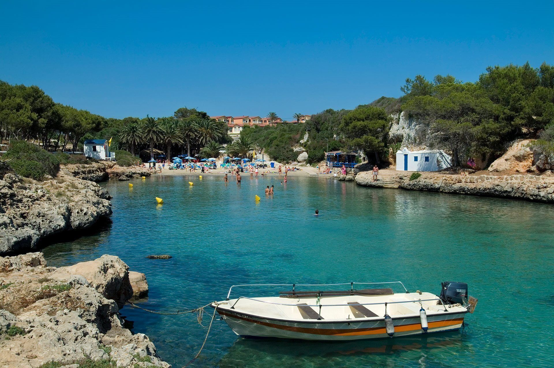 A coastal scene with a clear blue sea, a small boat docked near the shore, and a beach area with people and umbrellas.