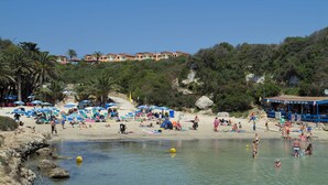 A beach with numerous umbrellas, a clear sky, and a building with a red roof in the background. - Villa Aljub by Villa Plus (Ciutadella de Menorca)