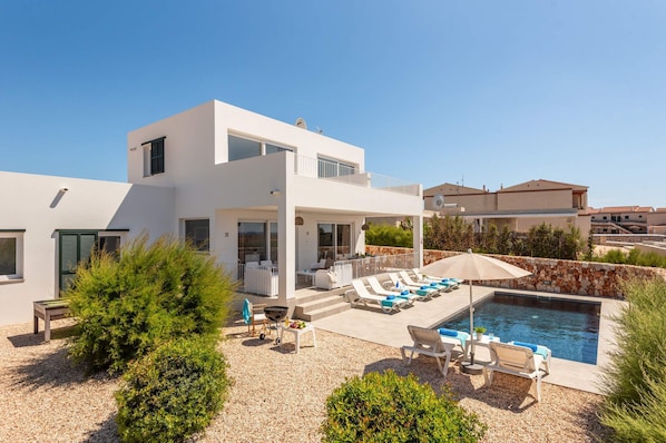 A modern two-story house with a pool, outdoor seating, and a stone wall.