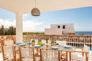 A terrace with a table set for dining, overlooking a pool and a modern building with a view of the sea.
