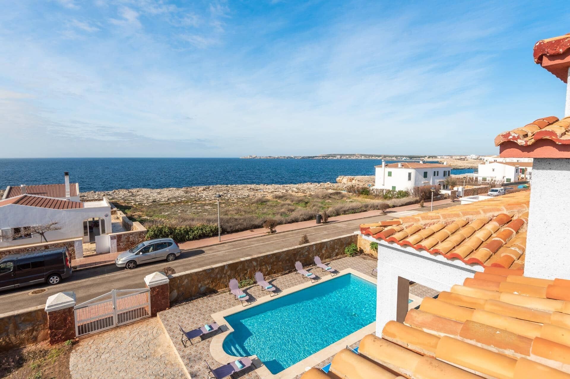 A pool area with a view of the sea, a road with vehicles, and buildings with red-tiled roofs.