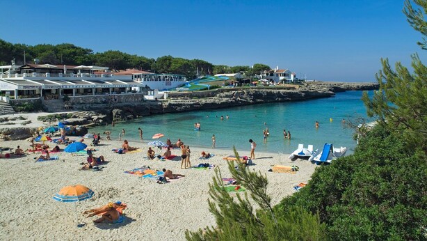 Ein Strand mit badenden Personen, einem Restaurant und einer felsigen Küstenlinie.
