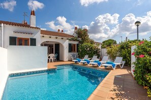 A pool area with lounge chairs and a small table, surrounded by a white building with a tiled roof and green shutters. - Villa Arena by Villa Plus (Ciutadella de Menorca)