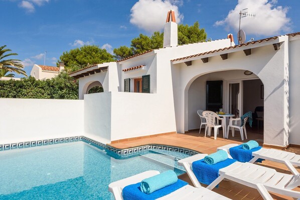 A pool area with lounge chairs and a table, surrounded by a white building with a tiled roof and a chimney.