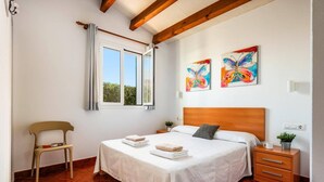 A bedroom with a bed, a nightstand, a chair, and two paintings on the wall. - Villa Avellano by Villa Plus (Cap d'Artrutx)
