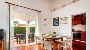 A dining area with a table set for four, a kitchen with wooden cabinets, and a sliding glass door leading to an outdoor patio with chairs. - Villa Avellano by Villa Plus (Cap d'Artrutx)
