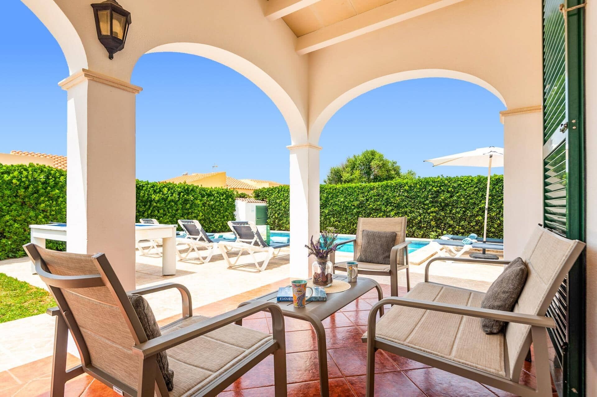 A covered outdoor seating area with lounge chairs and a table, overlooking a swimming pool and a hedge.