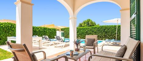 A covered outdoor seating area with lounge chairs and a table, overlooking a swimming pool and a hedge.