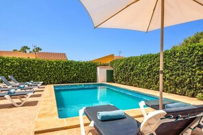 A swimming pool with lounge chairs and an umbrella, surrounded by hedges and buildings with red-tiled roofs.