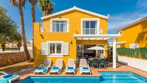 Una casa gialla a due piani con piscina, area relax all'aperto e palme.