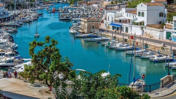 Una marina con numerose imbarcazioni ormeggiate lungo un canale stretto, circondata da edifici dall'architettura mediterranea.